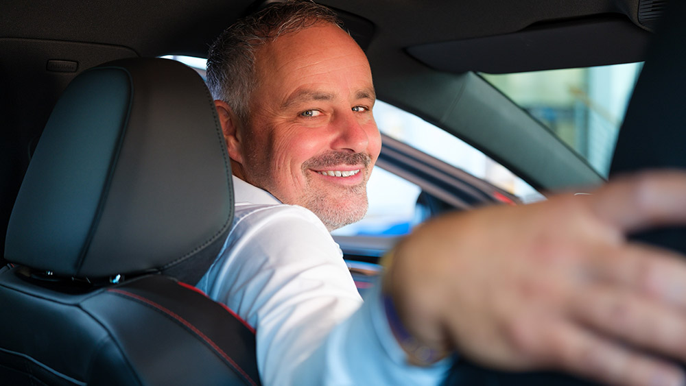 Mann sitzt im Auto, dreht sich lächelnd nach hinten und vermittelt Professionalität.