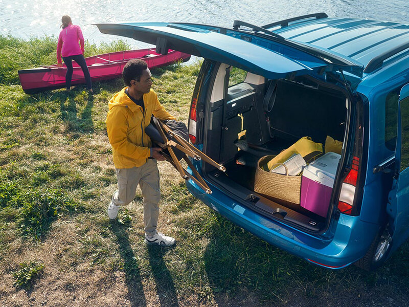 Ein blauer Volkswagen Caddy steht mit geöffneter Heckklappe an einem Gewässer. Eine Person lädt Sport- und Freizeit­ausrüstung in den großzügigen Laderaum, während im Hintergrund ein Kajak genutzt wird.