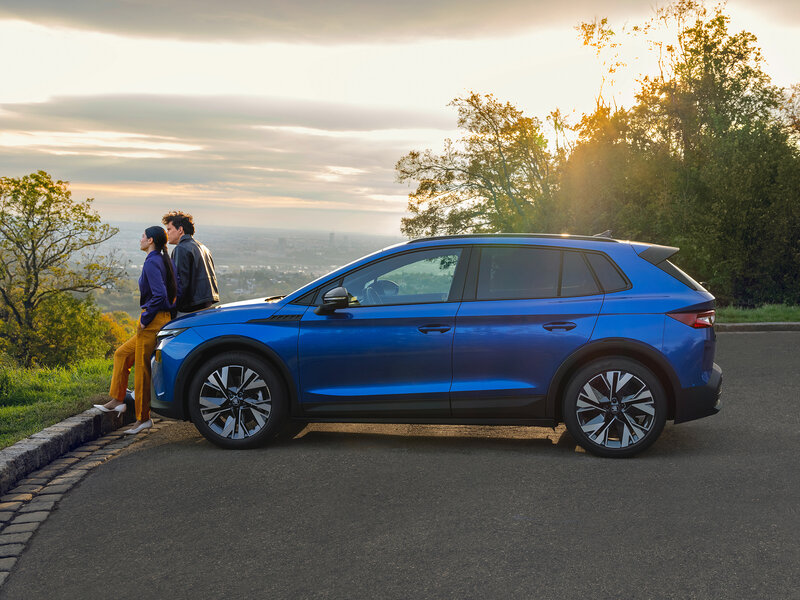Zwei Personen lehnen an einem blauen Škoda Elroq Sportline und genießen den Blick über die Landschaft bei Sonnenuntergang.