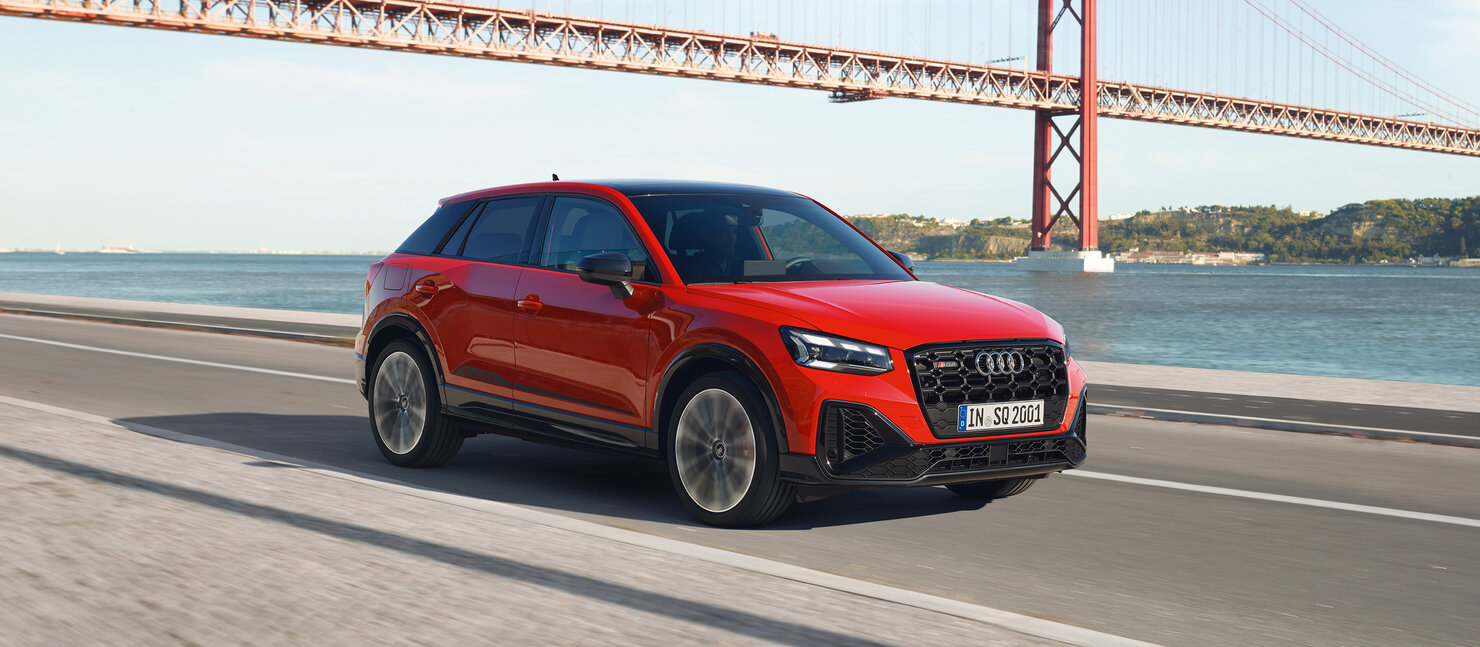 Audi SQ2 fährt auf Küstenstraße vor Brücke, rotes Performance-SUV mit dynamischer Front, großen Lufteinlässen und sportlichem Auftritt