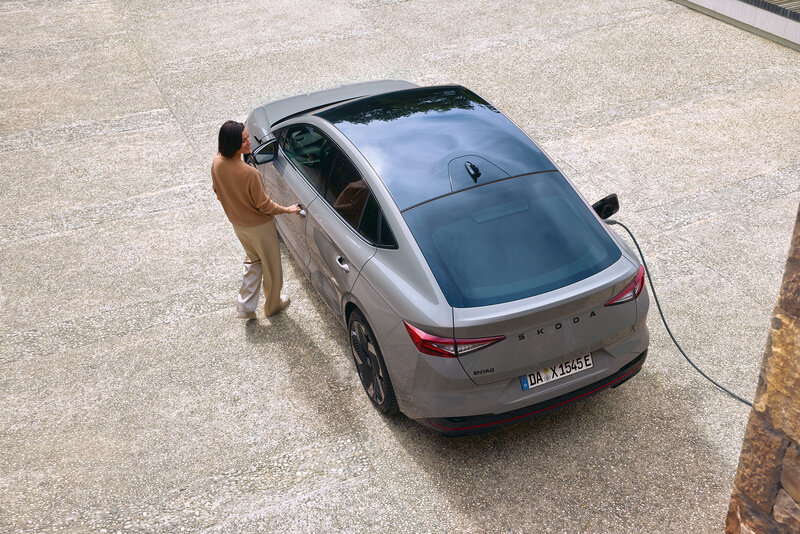 Eine Person lädt einen grauen Škoda Enyaq Coupé an einer Ladestation auf einem gepflasterten Platz.