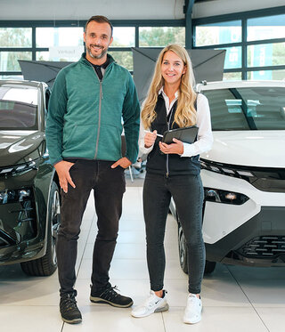 Zwei Servicemitarbeiter stehen im Autohaus zwischen zwei Fahrzeugen und lächeln in die Kamera.