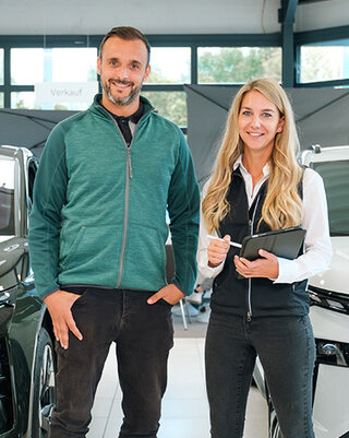 Zwei Servicemitarbeiter stehen im Autohaus zwischen zwei Fahrzeugen und lächeln in die Kamera.