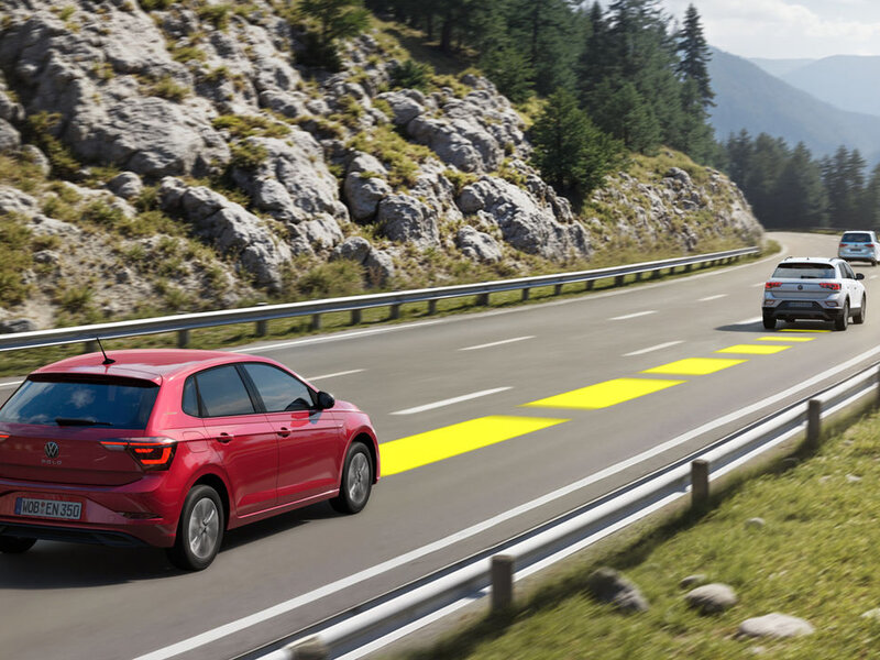 Ein roter Volkswagen Polo fährt auf einer Landstraße; grafische Markierungen visualisieren ein aktives Assistenzsystem.