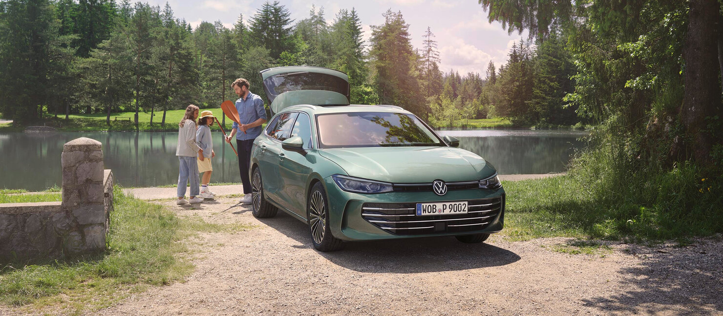 Grüner Volkswagen Passat steht mit geöffneter Heckklappe an einem See, während eine Familie Gepäck einlädt