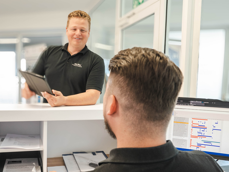 Zwei Mitarbeiter im Auto Bach Autohaus unterhalten sich an einer Empfangstheke mit Computer und Tablet im Bürobereich
