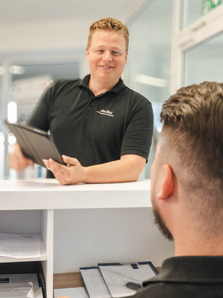 Zwei Mitarbeiter im Auto Bach Autohaus unterhalten sich an einer Empfangstheke mit Computer und Tablet im Bürobereich
