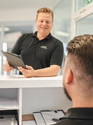 Zwei Mitarbeiter im Auto Bach Autohaus unterhalten sich an einer Empfangstheke mit Computer und Tablet im Bürobereich