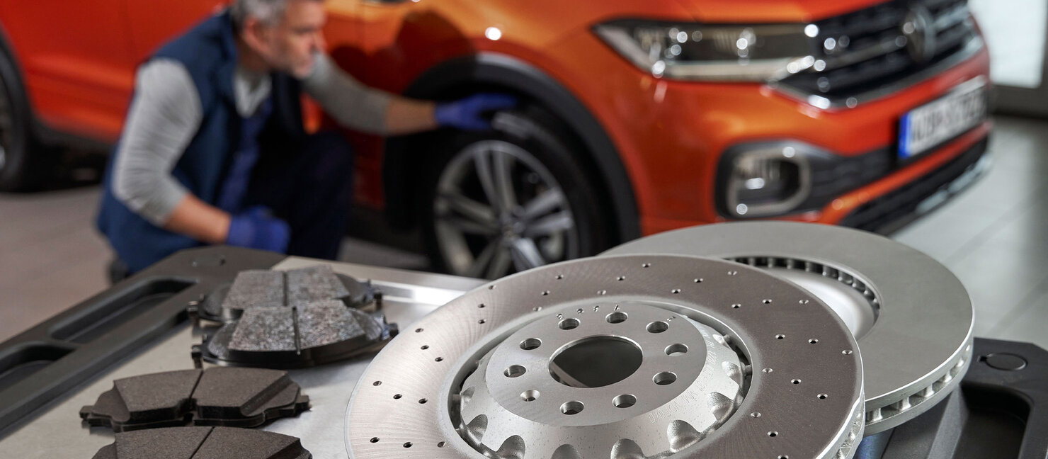 Bremsscheiben und Bremsbeläge liegen auf einem Werkstatttisch, im Hintergrund ein Mechaniker bei der Wartung eines orangefarbenen Volkswagen SUV