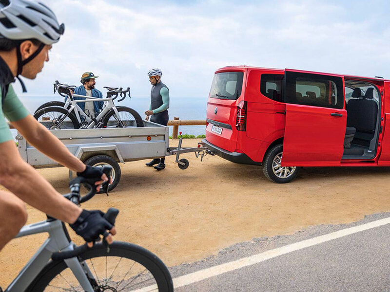 Ein roter Volkswagen Caravelle steht mit geöffneter Schiebetür auf einem Parkplatz. Mehrere Personen verladen Fahrräder auf einem Anhänger, Fokus auf Raumangebot und Transportflexibilität.