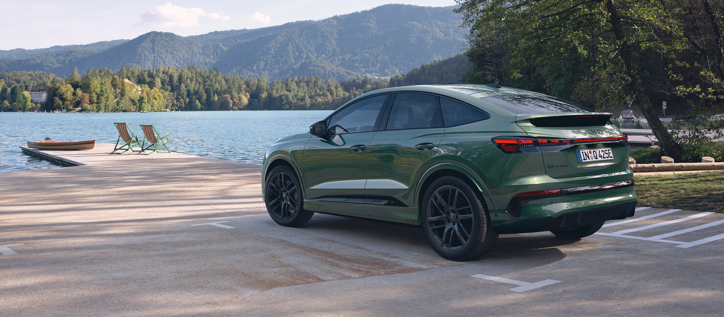Audi Q4 Sportback e-tron in Grün steht aus Heckperspektive auf Parkplatz an Seeufer mit Bergen und Bootsanleger im Hintergrund