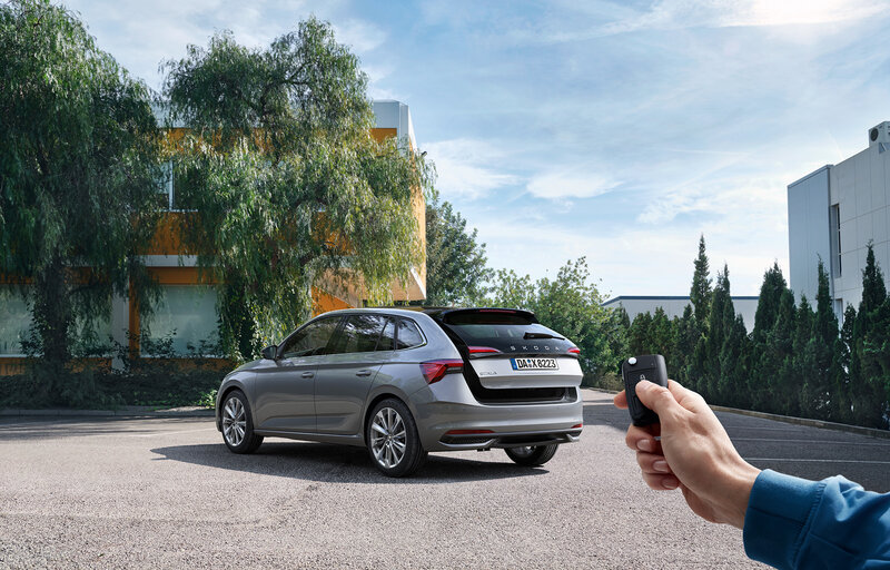 Eine Person öffnet per Funkschlüssel den Kofferraum eines grauen Škoda Scala auf einem Parkplatz vor Gebäuden