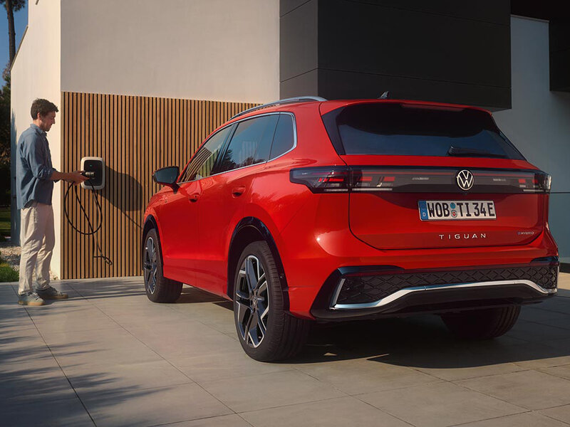 Ein roter Volkswagen Tiguan steht vor einem Gebäude und ist über ein Ladekabel mit einer Wallbox verbunden.