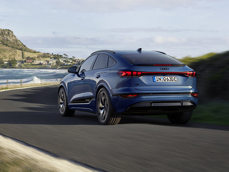 Audi Q6 Sportback e-tron fährt dynamisch auf Landstraße mit coupéhafter Dachlinie und sportlichem Heck