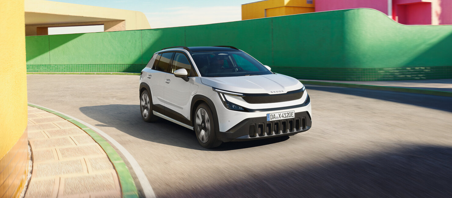 Škoda Epiq in Weiß fährt durch eine geschwungene Straße vor farbigen Gebäudefassaden in urbaner Umgebung