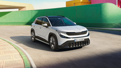 Škoda Epiq in Weiß fährt durch eine geschwungene Straße vor farbigen Gebäudefassaden in urbaner Umgebung