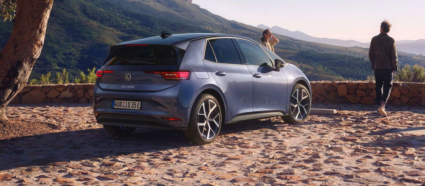 Heckansicht des Volkswagen ID.3 Neo geparkt auf gepflasterter Aussichtsterrasse mit zwei Personen und Blick auf bergige Landschaft bei Sonnenuntergang