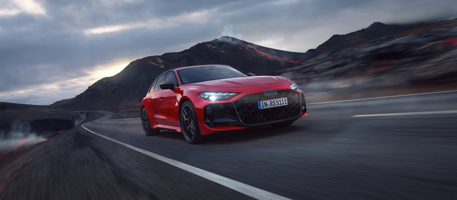 Audi RS5 Avant in Rot fährt dynamisch auf kurviger Landstraße vor Berglandschaft bei bewölktem Himmel