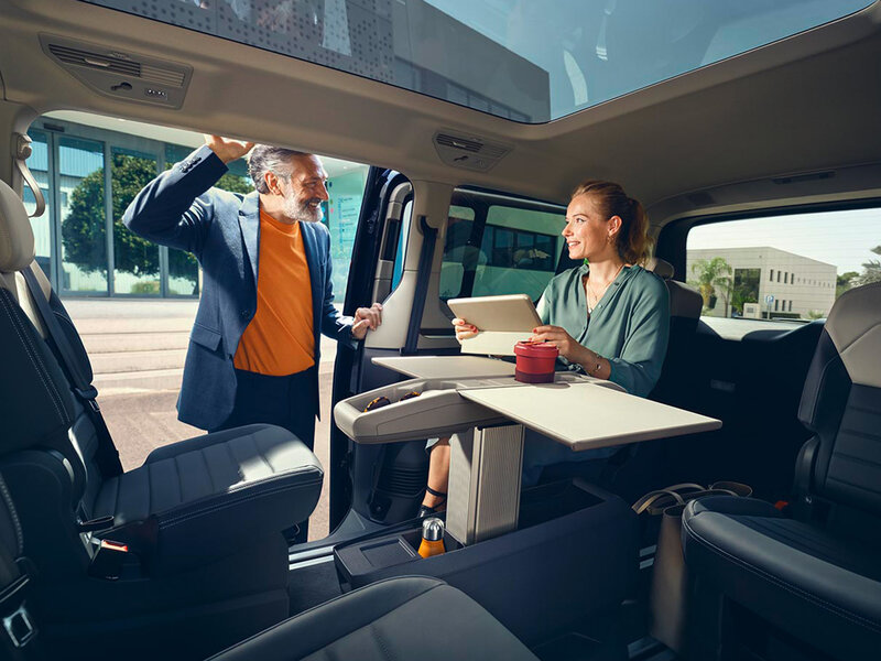 Innenraum des Volkswagen Multivan mit geöffneter Schiebetür. Zwei Personen stehen und sitzen im großzügigen Fahrgastraum, die flexible Sitzanordnung ist sichtbar.