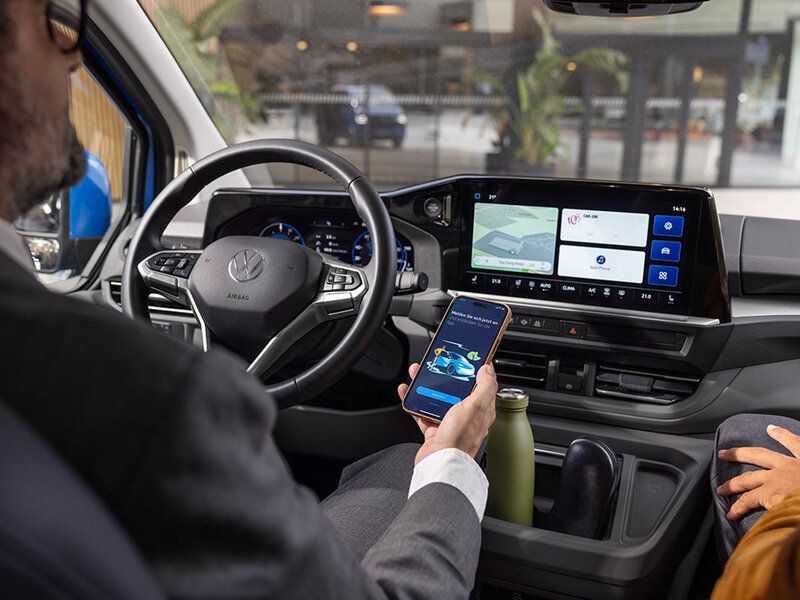 Blick auf den Fahrerplatz im Volkswagen Caravelle mit Multifunktionslenkrad, digitalem Cockpit und zentralem Infotainmentdisplay. Eine Person hält ein Smartphone zur Nutzung der Konnektivitätsfunktionen.