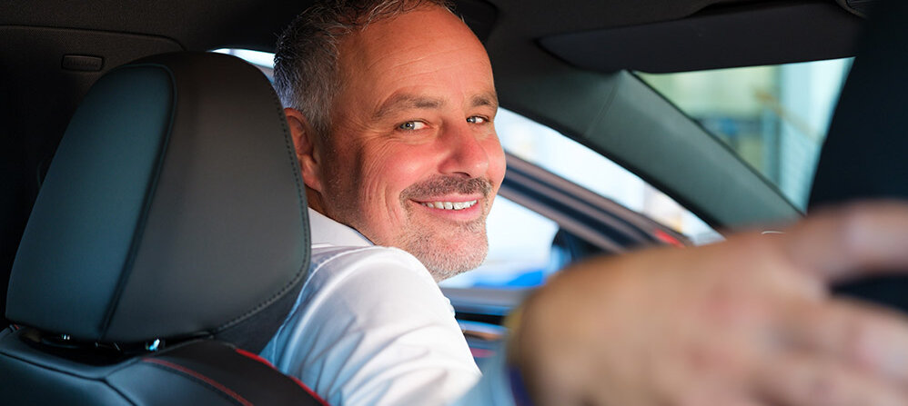 Mann sitzt im Auto, dreht sich lächelnd nach hinten und vermittelt Professionalität.