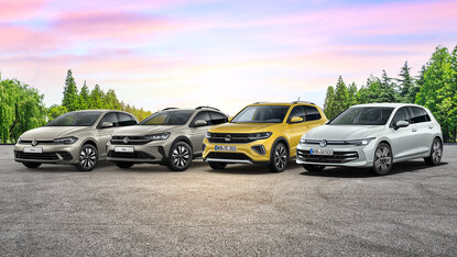 Vier Volkswagen Modelle stehen nebeneinander auf einer asphaltierten Fläche vor einer Baumreihe bei Sonnenuntergang