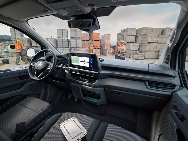 Blick in das Cockpit eines Volkswagen Transporters mit Multifunktionslenkrad, digitalem Display und Infotainmentsystem. Im Hintergrund ist eine Baustelle zu sehen.