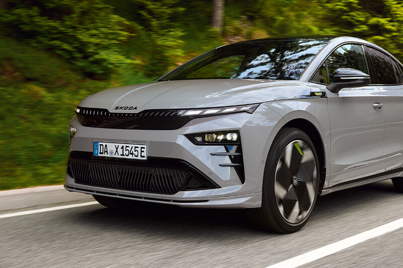 Škoda Enyaq Coupé in Stahl-Grau fährt auf einer kurvigen Straße durch bewaldete Landschaft.