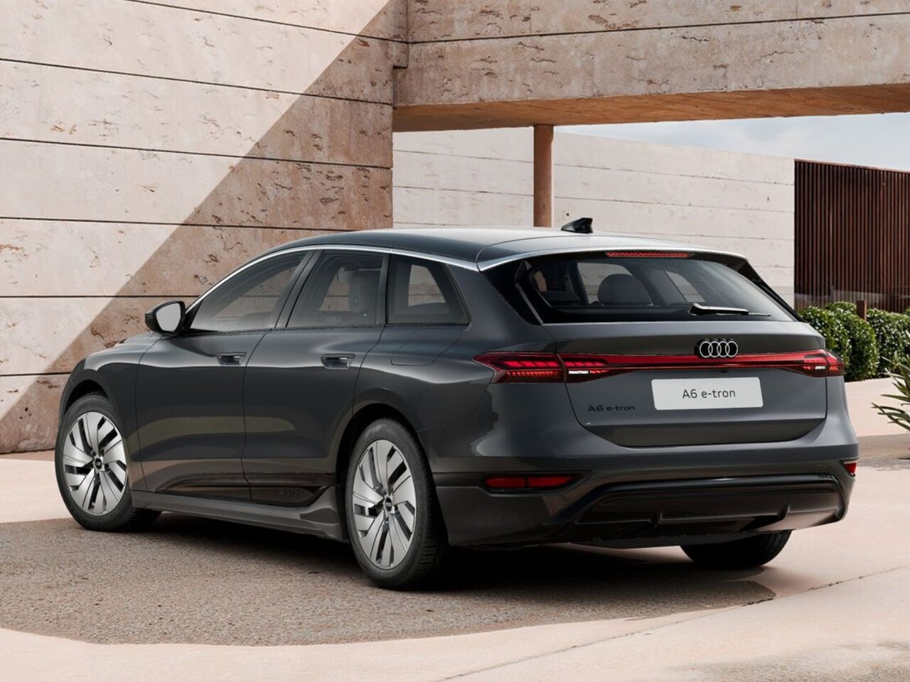 Dunkelgrauer Audi A6 Avant e-tron in Heckansicht steht vor moderner Architektur mit Steinfassade und begrüntem Außenbereich