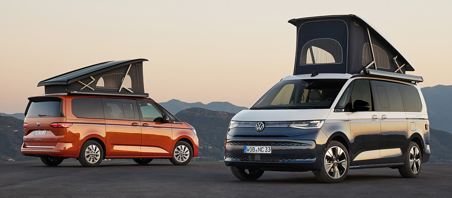 Zwei Volkswagen California stehen nebeneinander in einer offenen Landschaft bei Abendlicht. Ein Fahrzeug ist orange, das andere blau, beide haben ein geöffnetes Aufstelldach. Im Hintergrund sind Berge und ein klarer Himmel zu sehen.