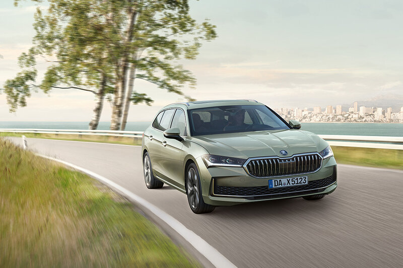 Škoda Superb Combi in Ice Tea-Beige Metallic, unterwegs auf einer kurvigen Küstenstraße mit Blick auf das Meer und eine entfernte Stadt.