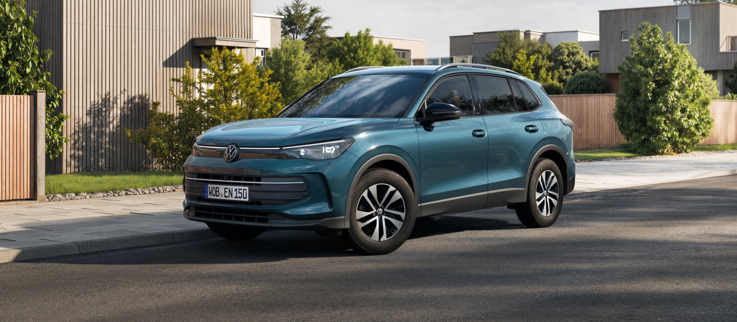 Volkswagen Tiguan in Petrolblau, Seiten-Front-Perspektive, fährt durch ruhiges Wohngebiet mit moderner Architektur