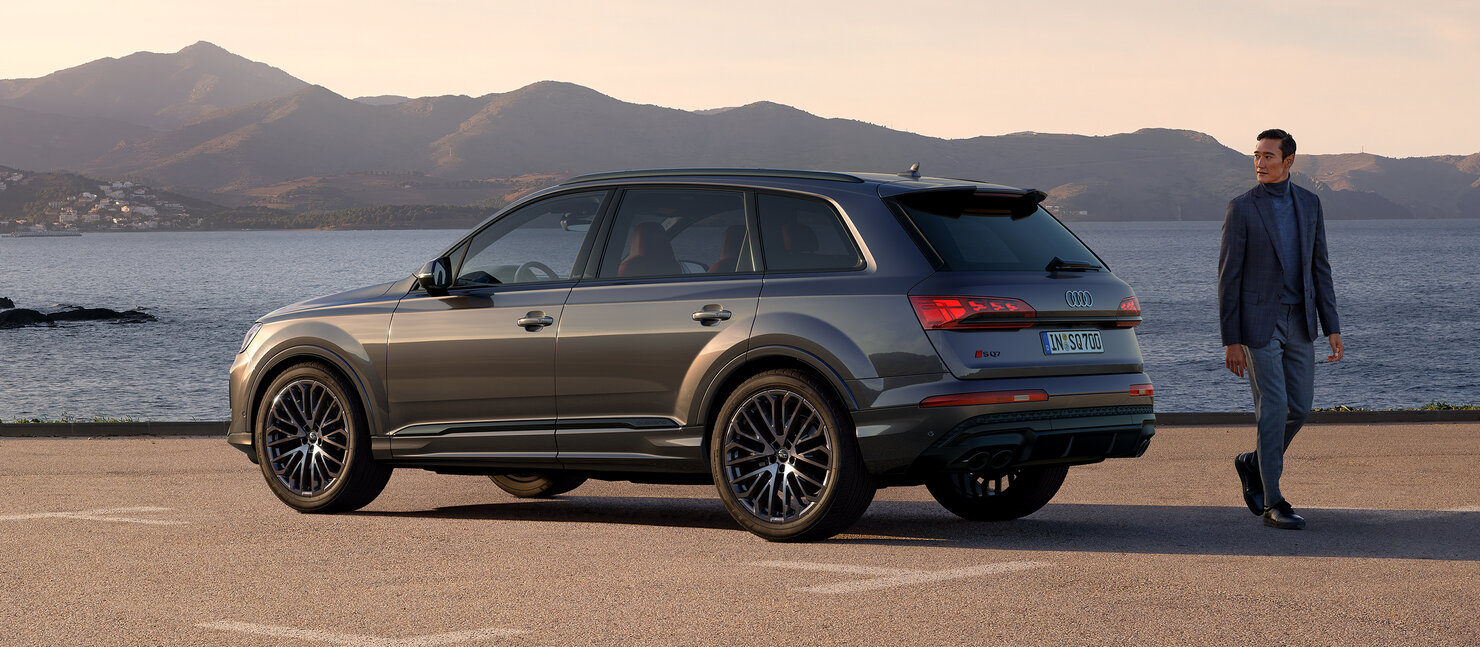 Audi SQ7 steht am Ufer vor ruhiger Bergkulisse, seitlich geparkt, daneben geht eine Person am Fahrzeug vorbei