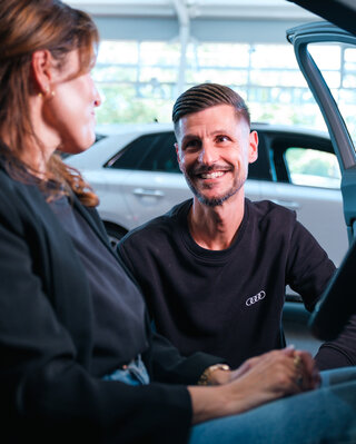 Verkaufsberater im Autohaus, der einer Kundin freundlich ein Fahrzeug erklärt, beide im Gespräch neben einem Audi-Modell.