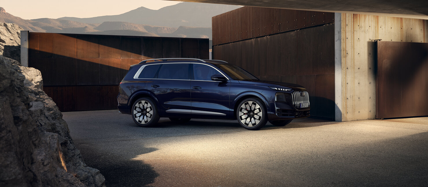 Audi Q9 in Dunkelblau aus Seitenperspektive vor moderner Wohnarchitektur mit Einfahrt und bergiger Landschaft im Hintergrund