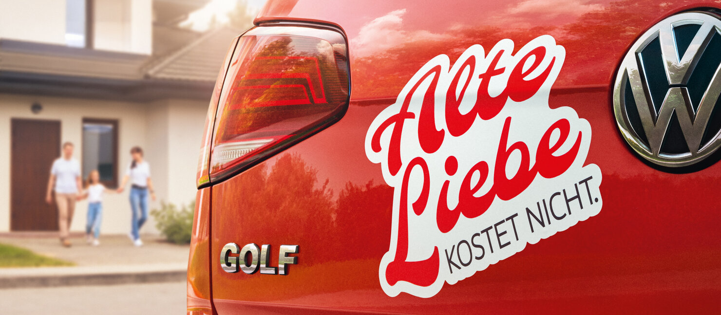 Roter Volkswagen Golf mit Aufkleber „Alte Liebe kostet nicht“ auf dem Heck, geparkt vor einem Haus mit einer Familie im Hintergrund