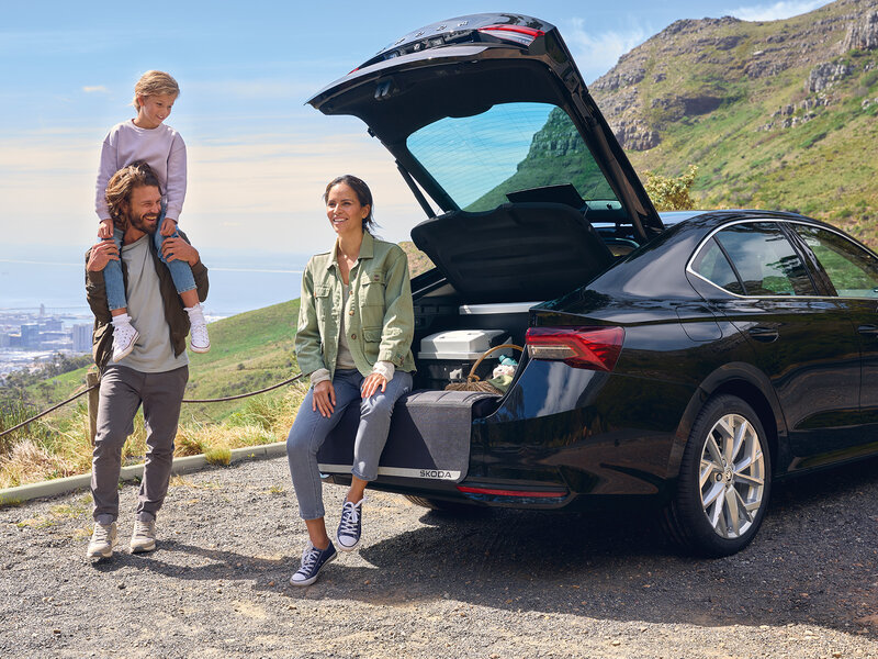 Drei Personen stehen neben einem schwarzen Škoda Octavia Limousine mit geöffnetem Kofferraum auf einer Bergstraße mit Blick ins Tal.
