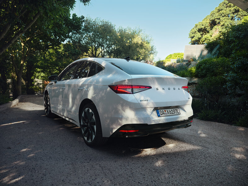 Škoda Enyaq Coupé in Weiß steht auf einer Straße im Grünen bei sonnigem Wetter.
