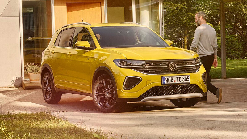 Gelber Volkswagen T-Cross steht in einer Einfahrt vor einem modernen Haus, eine Person geht am Fahrzeug vorbei