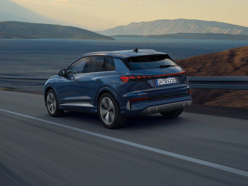 Audi Q4 e-tron in Blau fährt aus Heckperspektive auf Küstenstraße mit Meer und Bergen im Hintergrund