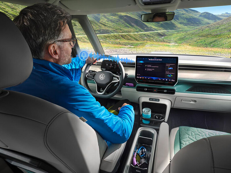 Blick aus dem Fond auf den Fahrerplatz des Volkswagen ID. Buzz während der Fahrt. Zu sehen sind das digitale Cockpit, das zentrale Touchdisplay und die Bedienung durch den Fahrer.