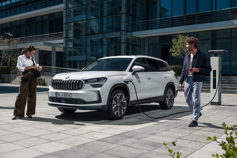 Zwei Personen stehen neben einem weißen Škoda Kodiaq iV, der an einer Ladestation vor einem modernen Bürogebäude lädt.
