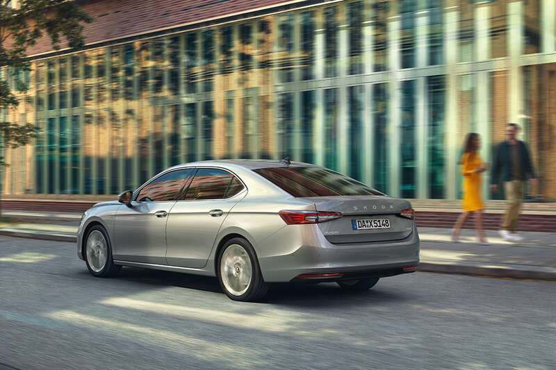 Auto Bach Alt-Text Generator sagte:  Škoda Superb Limousine in silberner Lackierung, unterwegs in einer modernen Stadt mit Glas- und Backsteinfassaden im Hintergrund