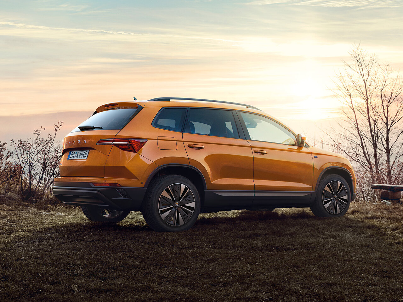 Škoda Karoq  in Phoenix-Orange Metallic steht auf einer Wiese bei Sonnenuntergang, im Hintergrund sitzen zwei Personen auf einer Bank.