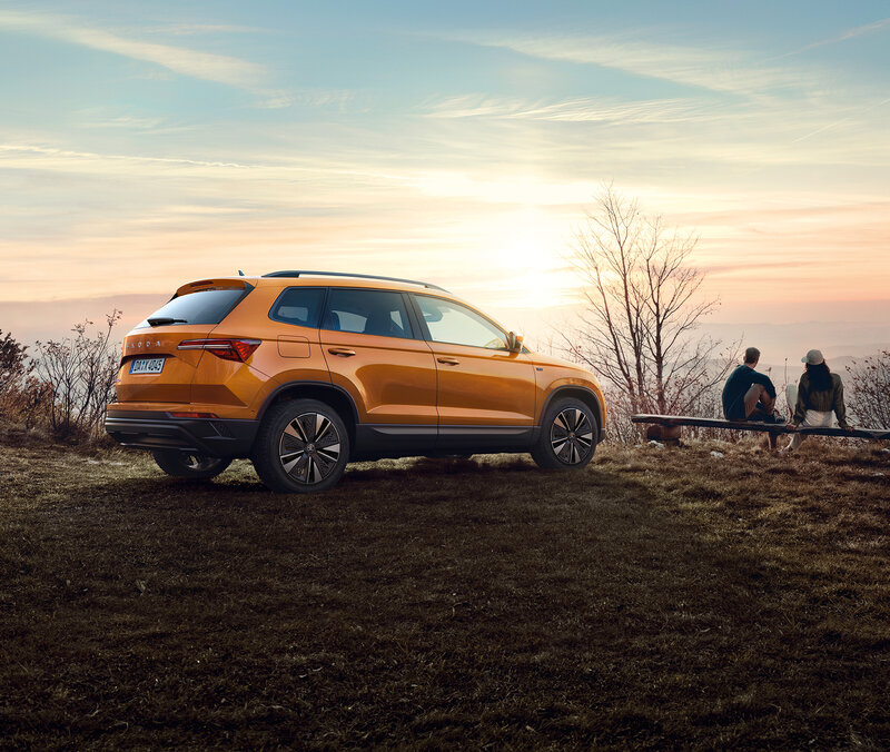 Škoda Karoq  in Phoenix-Orange Metallic steht auf einer Wiese bei Sonnenuntergang, im Hintergrund sitzen zwei Personen auf einer Bank.
