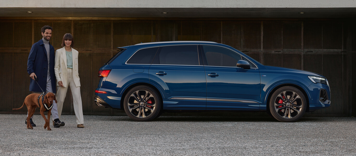 Blauer Audi Q7 steht seitlich vor einer Garage, ein Paar mit Hund geht vorbei und betont die Alltagstauglichkeit