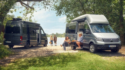 Silberner Volkswagen Grand California steht auf einem Parkplatz am Seeufer, während zwei Personen vor dem Camper an einem Tisch sitzen