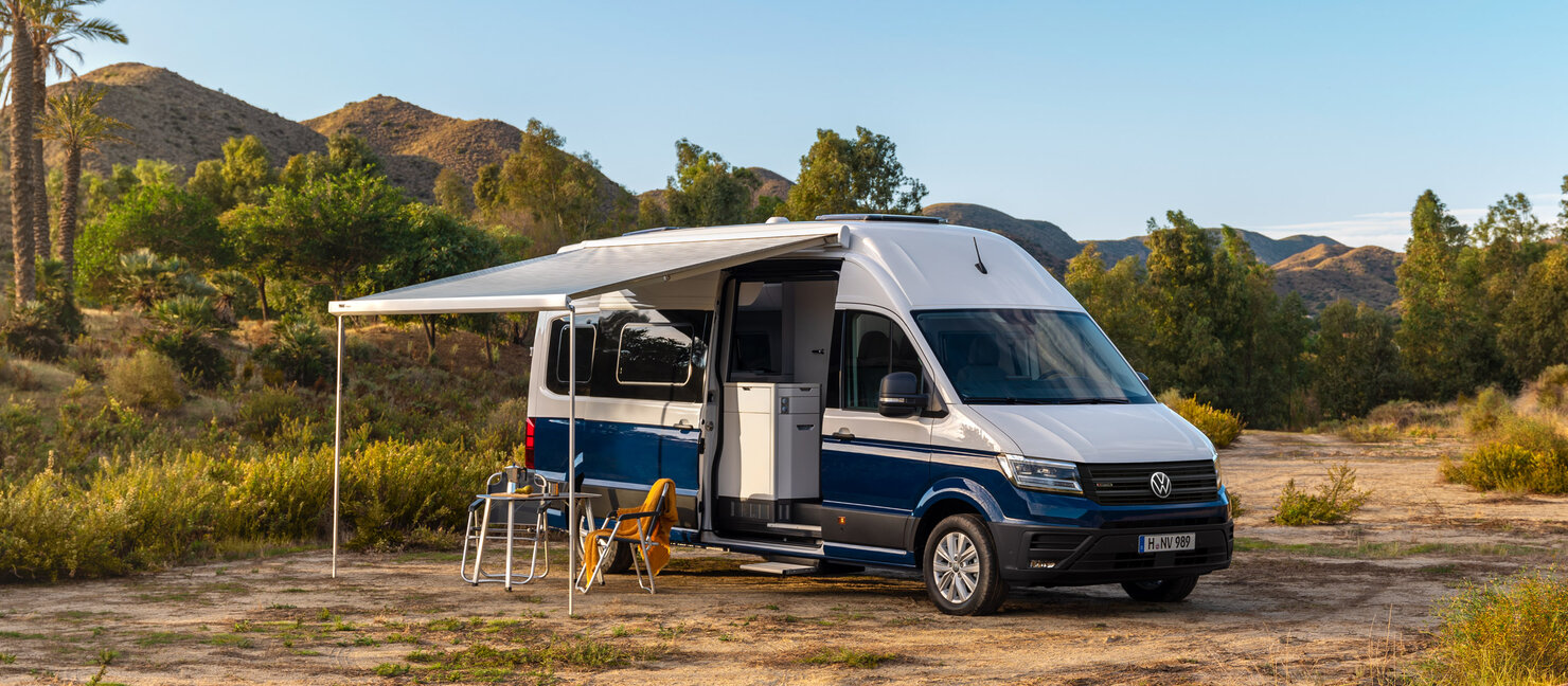 Ein weiß-blauer VW Grand California steht in einer Landschaft mit Bergen und Palmen. Die Markise des Campers ist ausgefahren, darunter stehen ein Tisch und zwei Stühle.