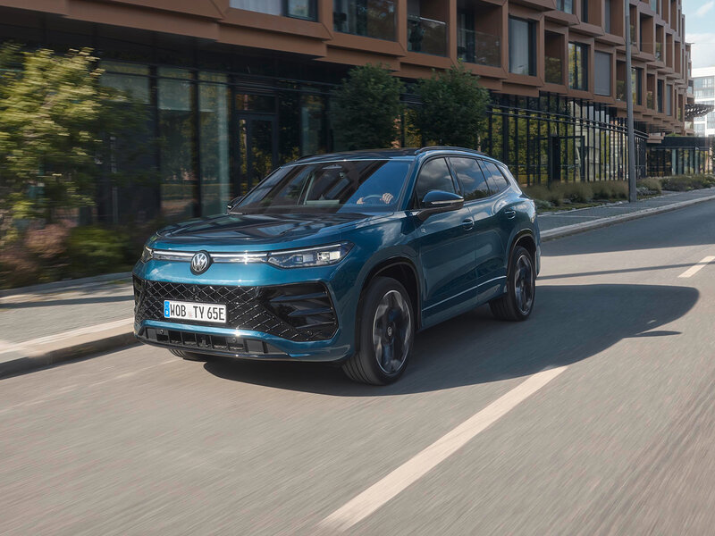 Volkswagen Tayron eHybrid in Blau fährt auf einer städtischen Straße vor moderner Gebäudefassade bei Tageslicht