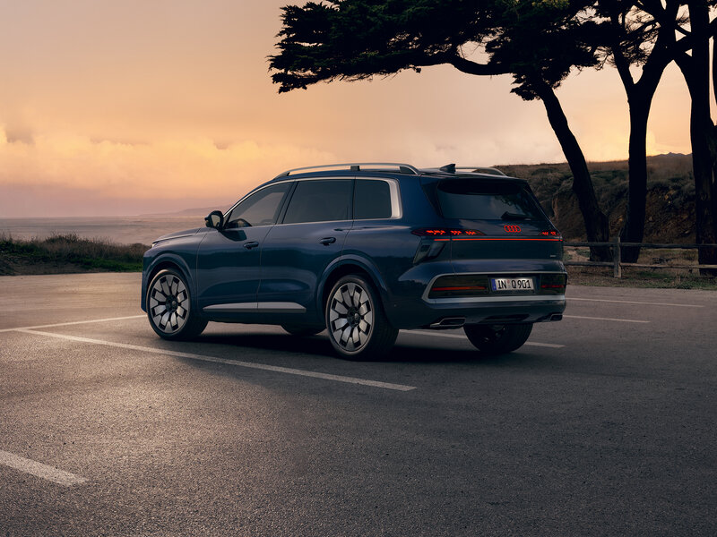 Audi Q9 in Dunkelblau aus Heckseitenperspektive auf einer Küstenstraße bei Sonnenuntergang mit Bäumen und Meer im Hintergrund